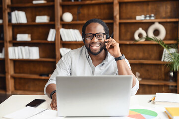 Cheerful African-American male employee wearing headset adjusts the microphone, holding a call, talking online with customers or coworkers, looks at the camera and smiles, using a laptop in the office