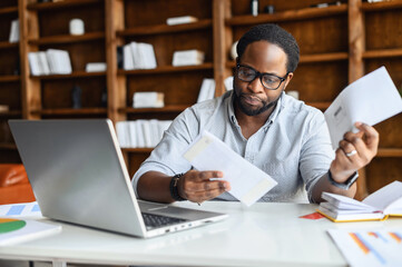 Worried African-American looking through correspondence, a guy holds envelopes in hands, got fine by post, concentrated man sitting at the desk with a laptop in contemporary office