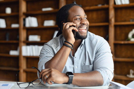 Cheerful African-American male employee wearing smart casual shirt holding phone conversation, a man sitting at the desk in modern office, a biracial entrepreneur talking over smartphone and laughs