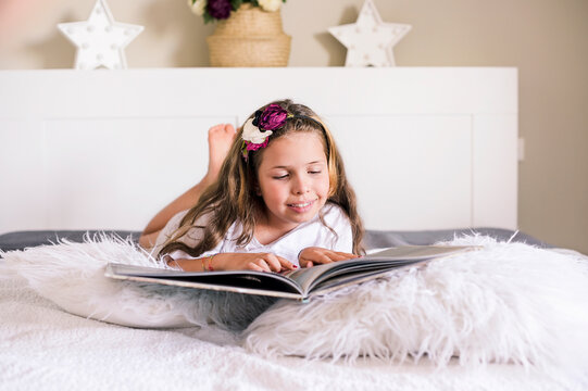 Little girl reads a book on the bed in a bright room. The child lies in the bedroom with a book and enthusiastically studies the book. Free space for text. High quality photo