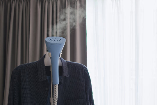 Electric garment iron steamer machine and dark blue shirt hanging on backdrop of blurred gray and white curtain with copy space, Wash and dry or Laundry, Ironing and hygiene sterilize with steam heat.