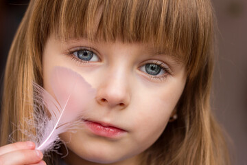 Close-up portrait of a little girl and a feather of a bird in her hands stroking her cheek, gentle...