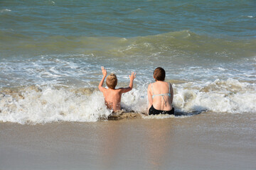 Two children sit in the sea and wait for the wave, boy with arms raised