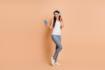 Fototapeta na wymiar Full length photo portrait of woman in headphones dancing holding phone in one hand isolated on pastel beige colored background