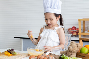 Little Caucasian girl wearing white chef hat cooking in kitchen. Child preparing easy food or bakery and lerning to cook by herselfe.