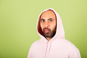 Young bald caucasian man in pink hoodie isolated on green background funny doing grimaces