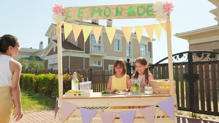 Medium shot of cheerful attractive woman with two elementary age girls getting prepared for outdoor market standing at wooden decorated diy stand with freshly made lemonade on hot summer day