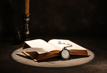 Open old book with clock, feather and candle on table against dark background