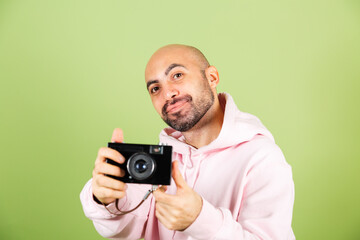 Young bald caucasian man in pink hoodie isolated on green background positive hipster hold camera
