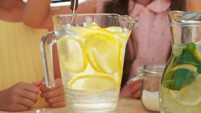 Midsection closeup of unrecognizable girls making lemonade outdoors in glass decanter stirring drink with spoon