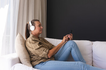 Happy caucasian senior woman in living room sitting on couch wearing headphones and using smartphone