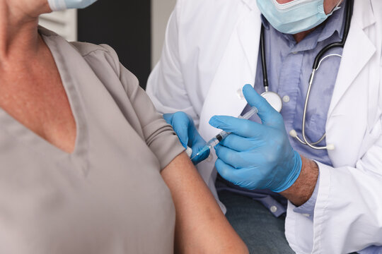 Midsection of caucasian male doctor wearing face mask giving female patient covid 19 vaccination