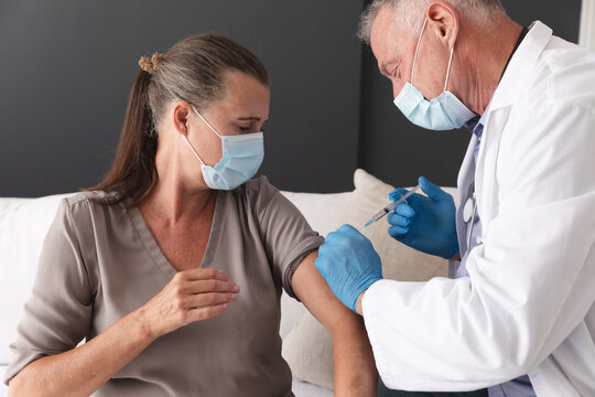 Caucasian Senior Male Doctor Giving Female Patient Covid 19 Vaccination, Both Wearing Face Masks