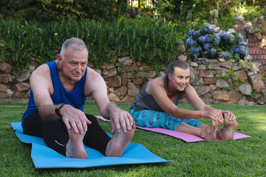 Happy caucasian senior couple exercising in garden, sitting on mats practicing yoga