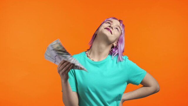 Happy violet haired woman with cash money - USD currency dollars banknotes on orange wall. Symbol of jackpot, gain, victory, winning the lottery