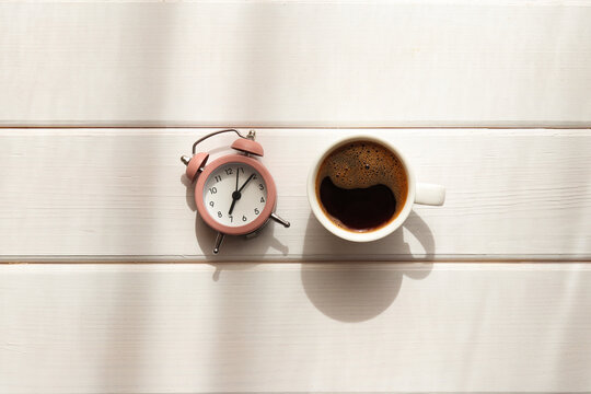 Cup of coffee and alarm clock on wooden table with shadows. Waking up, morning concept. Minimalism. Top view, flat lay