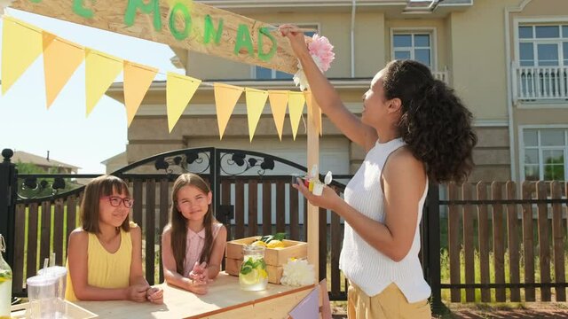 Dollying-in medium shot of joyful woman decorating wooden diy lemonade stand outdoors while two pretty elementary age girls waiting at counter smiling to camera, getting ready for market