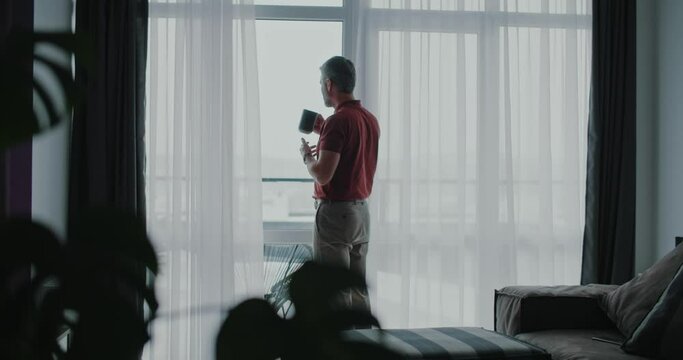 Shot back attractive man drinking coffee in the morning near window at home. Cozy atmosphere, windowsill, relaxation, standing, decor, design. Slow motion
