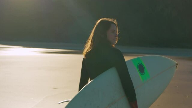 Slow motion shot camera follow beautiful authentic and real young woman in wetsuit walk with surfboard on sandy empty beach. Inspiring cinematic concept of candid female surfer