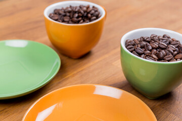 coffee composition, close-up of two cups of coffee one orange and one green with their plates full of coffee beans on a wooden surface, horizontal photo