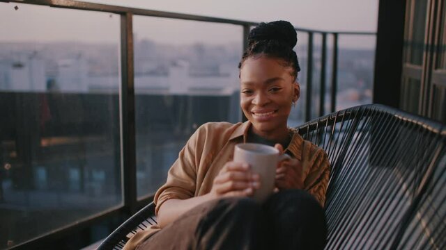 Happy young african american woman sitting on balcony looking out over city drinking a cup of tea or coffee. Relaxation,travel, interior, apartment. Slow motion