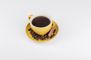 coffee composition, aerial shot of a yellow ceramic cup full of black coffee, on the coffee plate there are coffee beans and a cinnamon stick, the background surface is white, horizontal photo