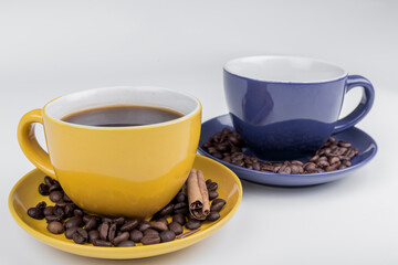 coffee composition, close-up of part of two ceramic cups, one yellow and one blue with their plates full of coffee beans and a piece of cinnamon stick on a white background, horizontal photo