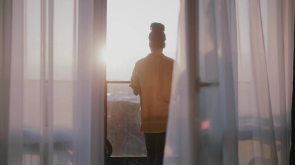 Attractive young african american woman stand on balcony looking out over city drinking a cup of tea or coffee. Relaxation,travel, interior, apartment. Slow motion - Powered by Adobe