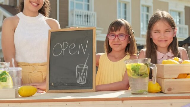 Handheld medium portrait of smiling attractive mixed race woman with two pretty elementary age daughters looking at camera standing at wooden stand selling homemade lemonade on hot summer day