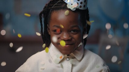 Crop view of cheerful girl smiling and looking to camera. Pretty afro american kid with dreadlocks in birthday hat rejoicing and posing while confetti falling Blurrred background.