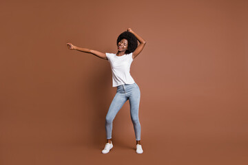 Fototapeta na wymiar Photo of carefree funny girl enjoy dance raise hands wear white t-shirt jeans footwear isolated brown color background
