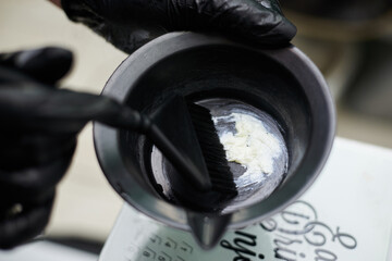 Close-up picture of hairdresser's tools in female hands with black gloves. Process of mixing hair dye with brush in plastic bowl. Hair stylist preparing for dying hair in barber shop. Beautification.