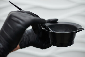 Close-up picture of hairdresser's tools in female hands with black gloves. Process of mixing hair dye with brush in plastic bowl. Hair stylist preparing for dying hair in barber shop. Beautification.