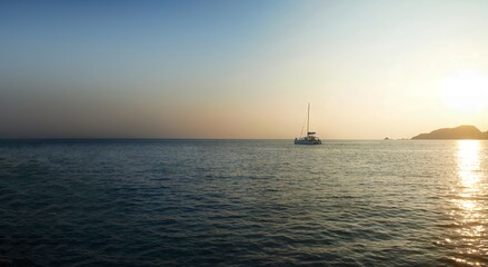 Horizontal panorama, space for text. Sailing yacht in the sea. Sunset, sun moves down over the rocky island. Reflection of the sparkling ray on a surface of water. Clear evening sky with colorful glow