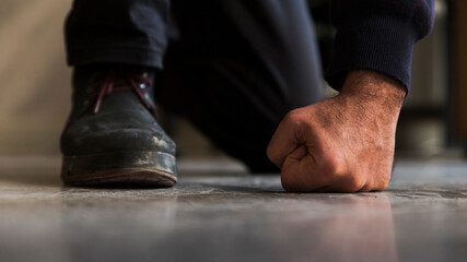 Close up detail of a crouched unrecognizable man's fist and foot with boot, standing on gray, screed floor in front shot.