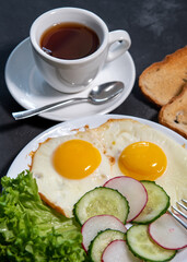 Two fried eggs, lettuce, cucumber and radish slices on a white plate, toast and a cup of tea or coffee. Scrambled eggs. Breakfast. Healthy eating. Close-up