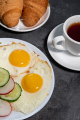 Two fried eggs, a leaf of lettuce, slices of cucumber and radish on a white plate, toast, croissants and a cup of tea or coffee. Breakfast. Healthy eating. Close-up