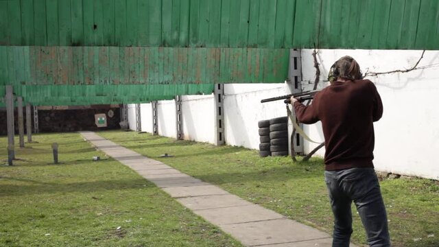 A young man shoots a pump-action firearm at flags, targets. A man in protective glasses and headphones in a dash. A green field with bullet protection outdoor. Sports shooting, weapons hobby. shotgun