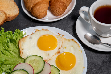 Two fried eggs, a leaf of lettuce, slices of cucumber and radish on a white plate, toast, croissants and a cup of tea or coffee. Breakfast. Healthy eating. Close-up