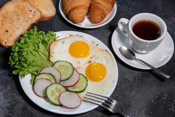 Two fried eggs, a leaf of lettuce, slices of cucumber and radish on a white plate, toast, croissants and a cup of tea or coffee. Breakfast. Healthy eating. Close-up