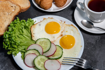 Two fried eggs, a leaf of lettuce, slices of cucumber and radish on a white plate, toast, croissants and a cup of tea or coffee. Breakfast. Healthy eating. Close-up