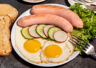 Two fried eggs and sausages, pieces of salad, cucumber and radish on a white plate, toast, croissants and a cup of tea or coffee. Scrambled eggs. Breakfast. Healthy eating. Close-up