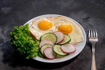 Two fried eggs with a leaf of lettuce, cucumber and radish slices on a white plate. Scrambled eggs. Breakfast. Healthy eating. Close-up