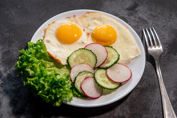 Two fried eggs with a leaf of lettuce, cucumber and radish slices on a white plate. Scrambled eggs. Breakfast. Healthy eating. Close-up