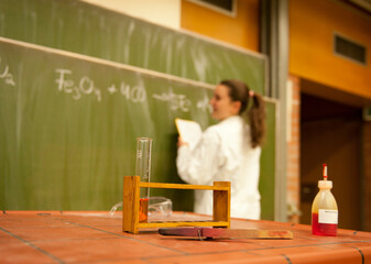 Junge Frau schreibt in einem Labor etwas auf eine Tafel, im Vordergrund steht ein Reagenzglas mit einer orangen Flüssigkeit