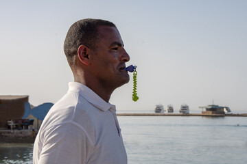 A dark-skinned man in his 40s blows a whistle against the backdrop of a seascape, close-up, selective focus Working as a lifeguard on the beach during the tourist season.
