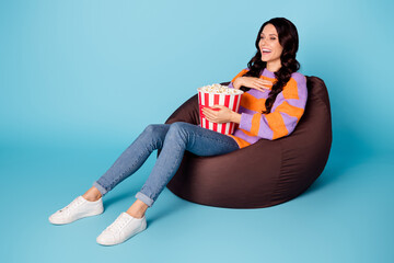 Portrait of pretty cheerful wavy-haired girl sitting on bag chair eating corn watching comedy...