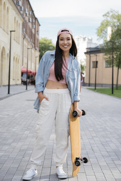 Portrait of young smiling teenager wearing stylish casual clothing standing on the street. Confident skater girl holding skateboard posing for pictures outdoors. Summer, positive lifestyle