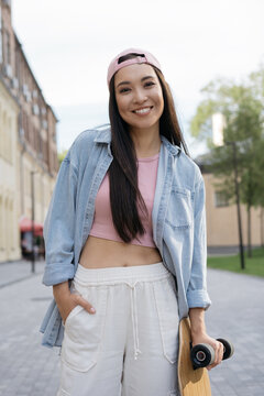 Portrait of beautiful asian woman wearing stylish casual clothing posing for pictures standing on the street. Young smiling teenager holding skateboard looking at camera outdoors. Positive lifestyle