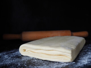 Raw puff pastry dough and wooden rolling pin on black background